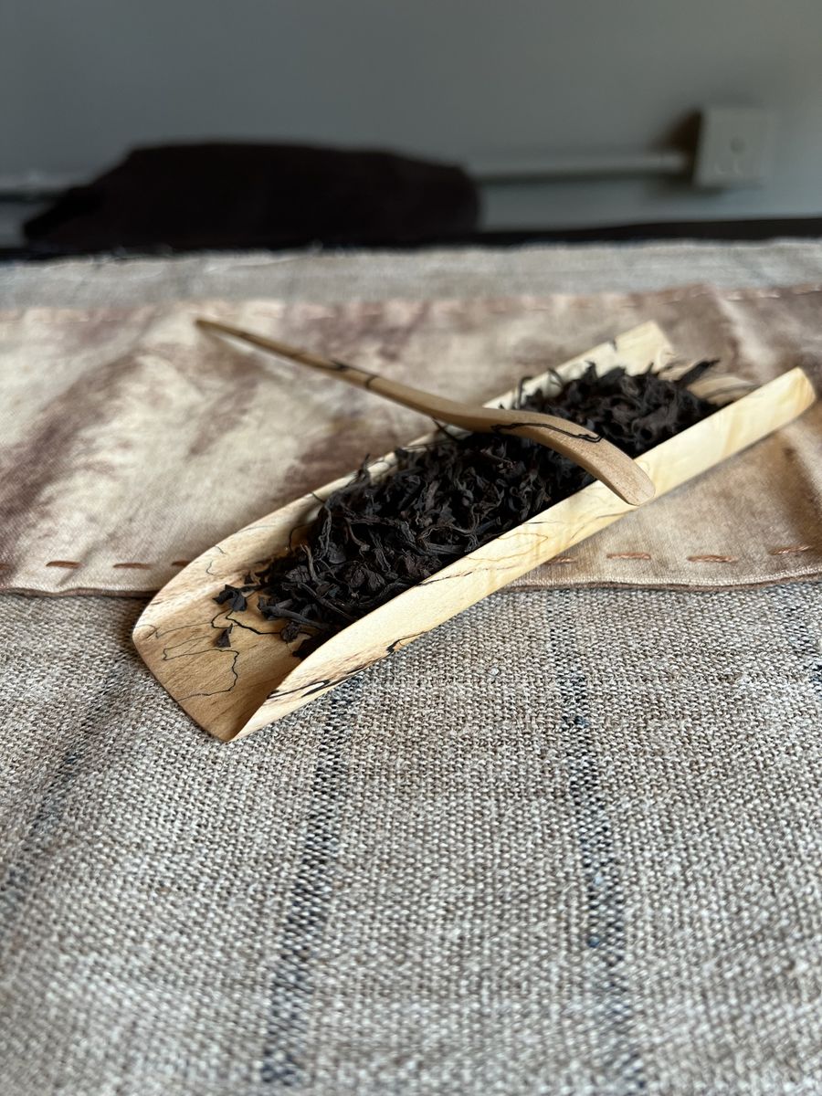 Dark twisted tea leaves resting in a pale wooden scoop on a linen cloth